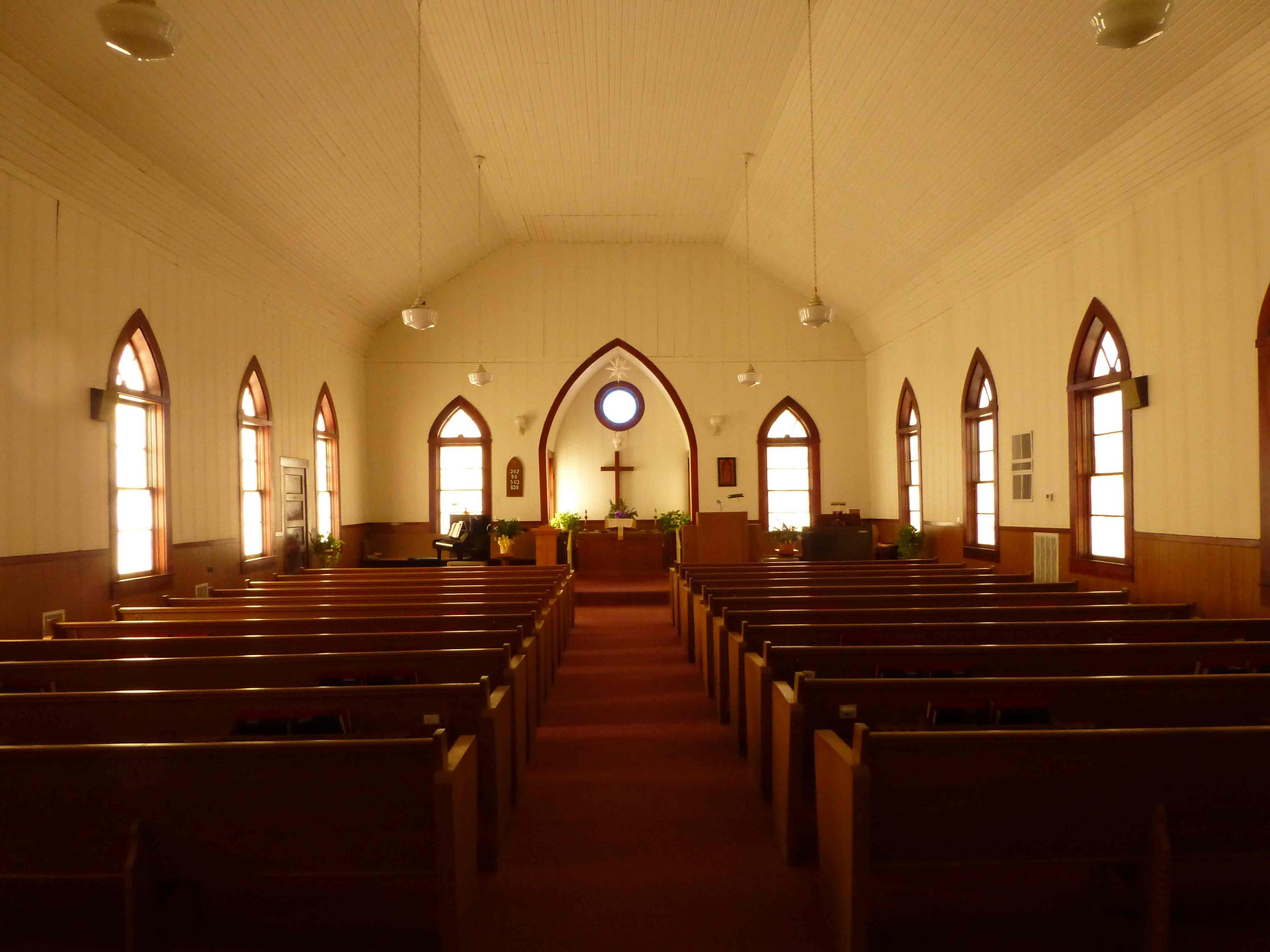 The sanctuary looking forward from the back pew: Gothic-arched windows on both sides, the chancel ahead, the pews warmed by late afternoon light.